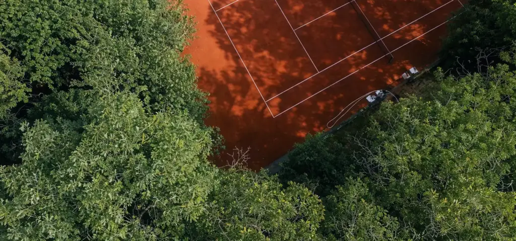 Clay tennis court design surrounded by dense green trees, viewed from above with shadows on the court