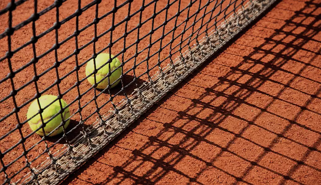 Tennis balls beside net on clay tennis court design with shadow patterns on the red surface