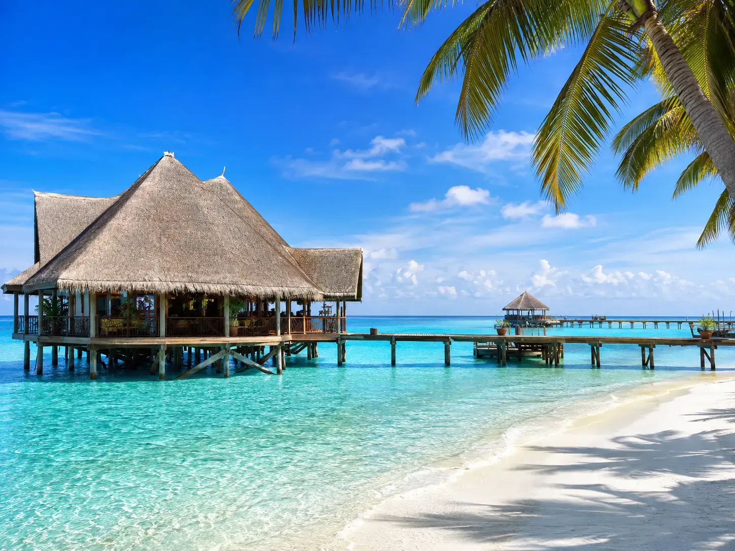 Overwater resort pavilion on stilts above turquoise lagoon with wooden pier, palm trees and white sand beach
