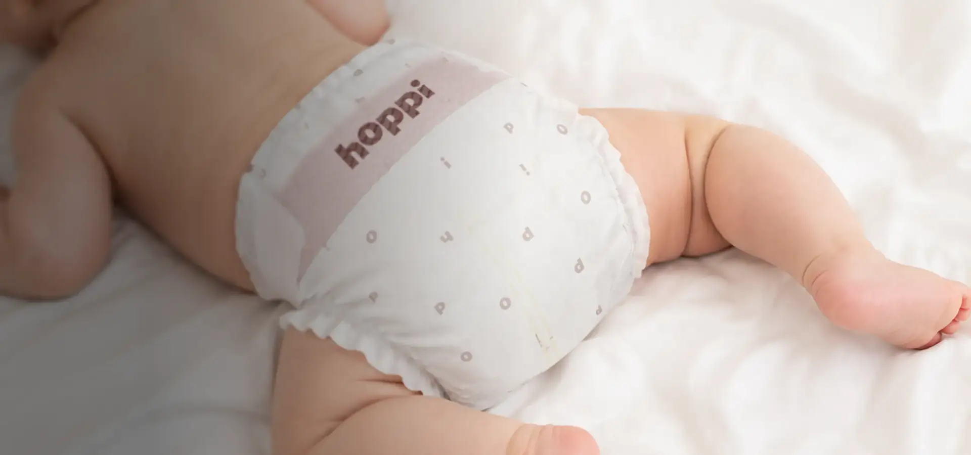 Baby lying on soft bedding wearing a hoppi diaper, baby diaper product visualization.